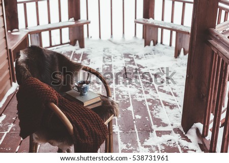 Chair with fur cover on a porch deck of a log cabin with snow. Tea, warm blanket and reading. Cold winter relax weekend