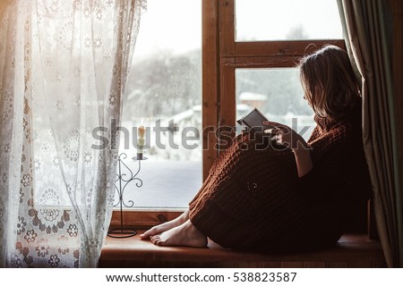 Pre teen child in warm woolen sweater seating on window sill and reading a book. Winter weekends in old log house. Cold snowy weather. Cozy homely concept.