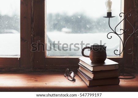 Warm and comfy winter concept. Book, cup of tea and candlestick on wooden window sill in old house. Reading and relaxing in cold snowy weather at home. Quiet silent homely scene.