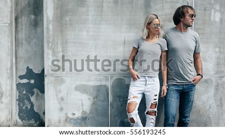 Two hipster models man and woman wearing blank gray t-shirt, jeans and sunglasses posing against rough concrete wall in the city street, front tshirt mockup for couple, urban style
