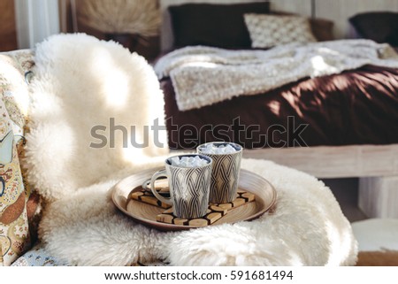 Still life details of nordic interior. Two cups of cocoa with marshmallows on wooden tray on a armchair with sheep fur cover in bedroom. Lazy winter morning in log cabin.