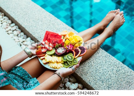 Girl relaxing and eating fruit plate by the hotel pool. Exotic summer diet. Photo of legs with healthy food by the poolside. Tropical beach lifestyle.