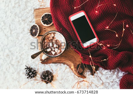 Winter homely scene. Warm knit blanket, phone with blank screen and cup of sweet cocoa with marshmallows on wooden tray in bed. Lazy cold weekend.