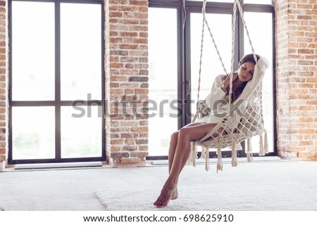 Young woman chilling at home in comfortable hanging chair in front of big window. Girl relaxing in swing in loft living room with brick walls. Beautiful legs barefoot on white carpet.