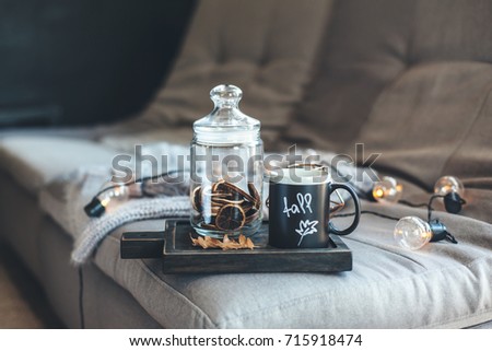Still life details of living room. Cup of coffee on rustic wooden tray, candle and warm woolen sweater on sofa, decorated with led lights. Autumn weekend concept. Fall home decoration.