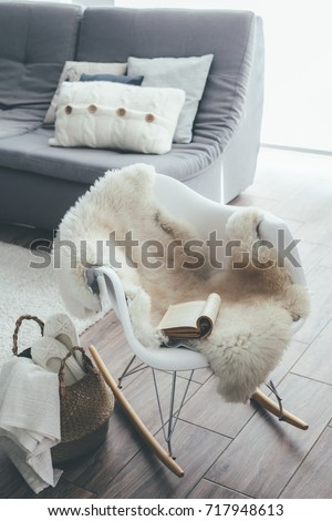 Still life details of living room. Sheep skin rug on modern armchair by the sofa with cushions. Reading book on the rocket chair. Cozy winter scene in Scandinavian interior.