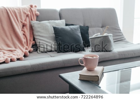 Cocoa with marshmallows and book and knitted basket with yarn and needles on coffee table. Warm pompon blanket and cushions on the sofa. Still life photo of nordic interior details.