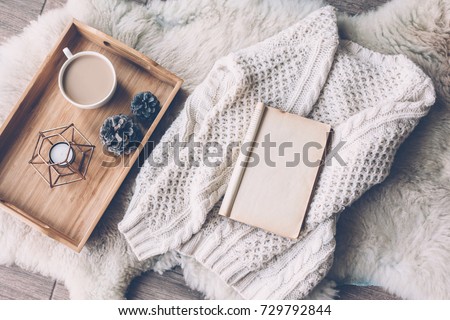 Mug with coffee and home decor on wooden serving tray on sheep skin rug. Warm sweater and open book, winter weekend concept, top view