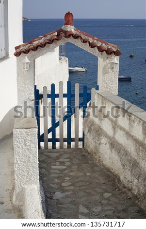 Stone-path leading to white and blue wooden gate with an arch and sea with boats behind the gate on a sunny day. Greek island.