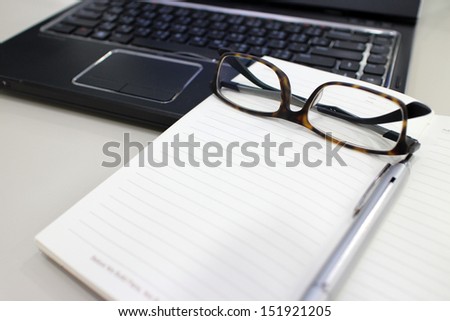 An office desk with office items in a work place scene there are workbooks on table and modern laptop, pen and glasses near on a gray background.