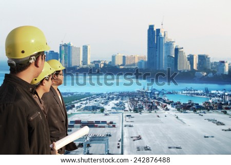 three engineer team standing in front of cranes at the container port terminal with big city background