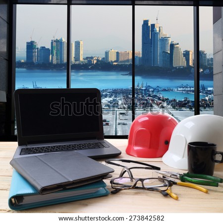 computer and builder\'s work tool on wooden table with cranes at the container port terminal with big city background