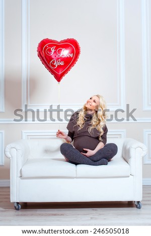 young beautiful pregnant woman sitting on sofa and looking at the balloon in the form of heart. Valentine\'s Day.