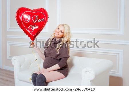 young beautiful pregnant woman sitting on a sofa and holding balloon in the form of heart in hand. Valentine\'s Day.