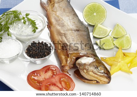 A smoked trout with salt, black pepper, tomato slices, lime pieces, a starfruit slice, dip, parsley and dill.