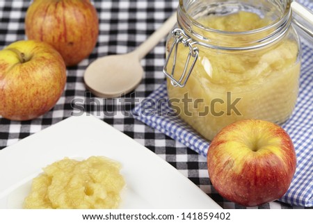 Apples, a wooden spoon and a bottling jar of stewed apples a ready on a black and white checkered tablecloth.