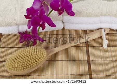 A bath brush is laying on a mat of bamboo. In the background are two pile of folded towels decorated with a purple orchid.
