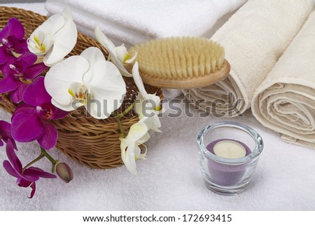 A bath brush with purple and white orchids into a basket standing in front of some rolled towels. The scene is decorated with a tealight into a glass bowl.