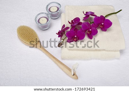 A bath brush is laying beneath two folded, naturally colored towels. The decoration is made of a purple orchid and two tealights into a glass bowl.