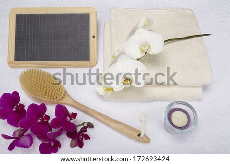 A bath brush is laying beneath two folded, naturally colored towels. A slate can be used as a free text area. The scene is decorated with a purple and white orchid and a tealight into a glass bowl.