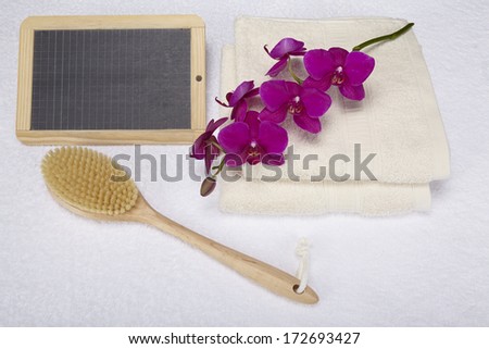 A bath brush is laying beneath two folded, naturally colored towels. A slate can be used as a free text area. The scene is decorated with a purple orchid.