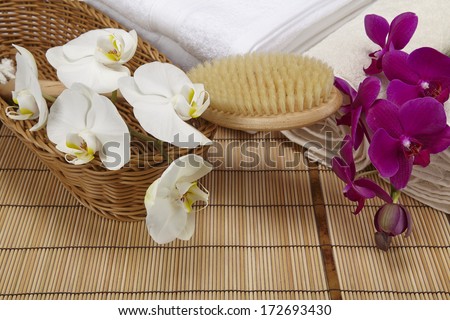 A bath brush with purple and white orchids into a basket standing in front of some rolled towels and a pile of folded towels. All objects are standing on a mat of bamboo.