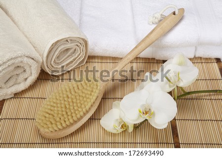 A bath brush is laying on a pile of white, folded towels. Beneath are two naturally colored, rolled towels and a white orchid.