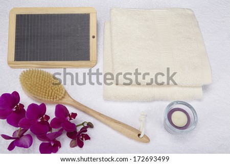 A bath brush is laying beneath two folded, naturally colored towels. A slate can be used as a free text area. The scene is decorated with a purple orchid and a tealight into a glass bowl.