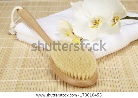 A bath brush is laying on a white folded towel. The scene is taken on a bamboo mat and decorated with a white orchid.
