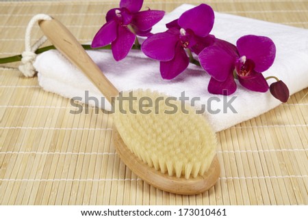 A bath brush is laying on a white folded towel. The scene is taken on a bamboo mat and decorated with a purple orchid.