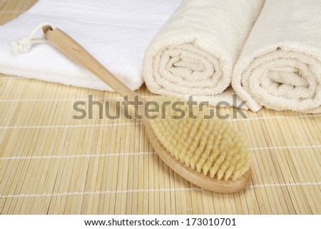 A bath brush is laying on a folded white towel on top of a bamboo mat. Beneath are two rolled naturally colored terry clothed towels.