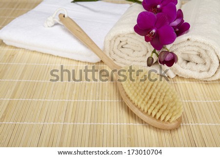 A bath brush is laying on a folded white towel on top of a bamboo mat. Beneath are two rolled naturally colored terry clothed towels. The scene is decorated with a purple orchid.