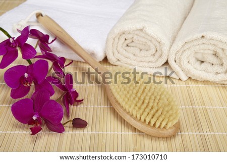 A bath brush is laying on a folded white towel on top of a bamboo mat. Beneath are two rolled naturally colored terry clothed towels. The scene is decorated with a purple orchid.