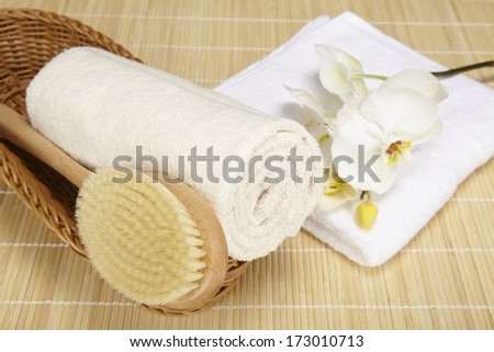 A bath brush and a rolled up terry clothed towel are laying into a basket. A folded white towel and an orchid are located beneath the basket. The objects are standing on a bamboo mat (bath mat).
