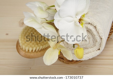 A bath brush and a rolled up terry clothed towel are laying into a basket. The basket stands on a tabletop of spruce wood and is decorated with a white orchid.