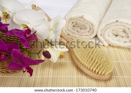A bath brush is laying on a folded white towel on top of a bamboo mat. Beneath are two rolled naturally colored terry clothed towels. The scene is decorated with a purple and white orchid.