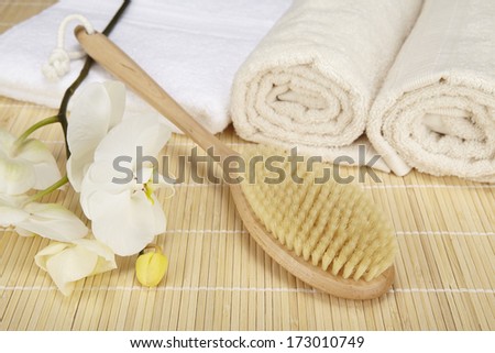 A bath brush is laying on a folded white towel on top of a bamboo mat. Beneath are two rolled naturally colored terry clothed towels. The scene is decorated with a white orchid.