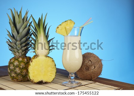 A Pina Colada, a half of a pineapple and a coconut are standiing on a tabletop of acacia wood. The background is blue.