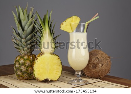 A Pina Colada, a half of a pineapple and a coconut are standiing on a tabletop of acacia wood. The background is gray.