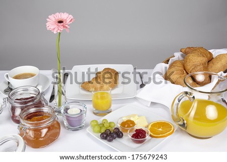A well-laid breakfast table with a cup of coffee, a basket of croissants and bread rolls, strawberry and apricot jam and decoration of a tealight and a rose gerbera