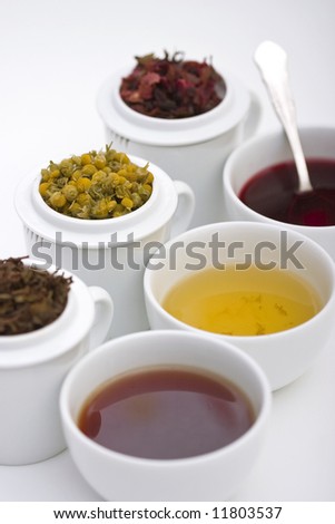 Three cups of tea in different colors with three different types of herbs in pot behind it