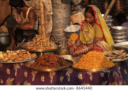 Woman in brightly colored sari selling indian sweets from a market stall.