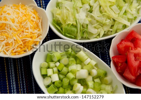 Veggie Dishes Small white bowls filled with cheese, lettuce, tomato, and scallions.  Close-up of the bowls of sliced vegetables and cheese.  The background is dark blue woven material.