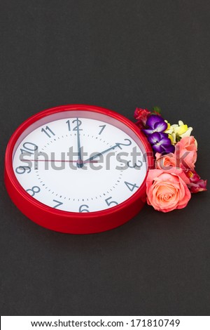 Daylight Saving Time Spring time daylight saving clock and floral arrangement.  The clock is red with black numbers, there is a spring floral arrangement next to the clock, centered at right side.