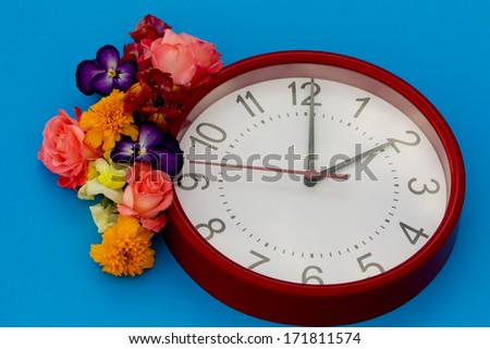 Daylight Saving Spring Red clock with black numbers on a blue back ground. At the upper left side of the clock is a spring floral arrangement. The clock is set at 2 am.