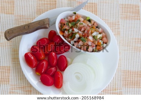 Making Pico De Gallo A small bowl of Pico De Gallo, on a white plate with grape tomatoes and onion. The plate is on a woven mat with a knife to the upper left side.