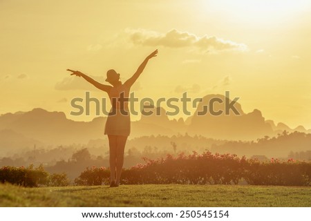 Woman\'s silhouette is standing on background of sunset sky and mountains and pulls her hands to the sun