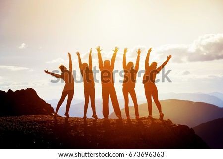 Five happy friends having fun standing with raised arms on mountain top at sunset time