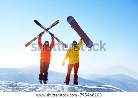 Two active friends skier and snowboarder are standing on mountain top against blue sky and mountains. Ski resort concept