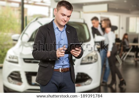 Let me assist you in your vehicle search. Handsome young classic  salesman standing at the dealership with the customers on the background